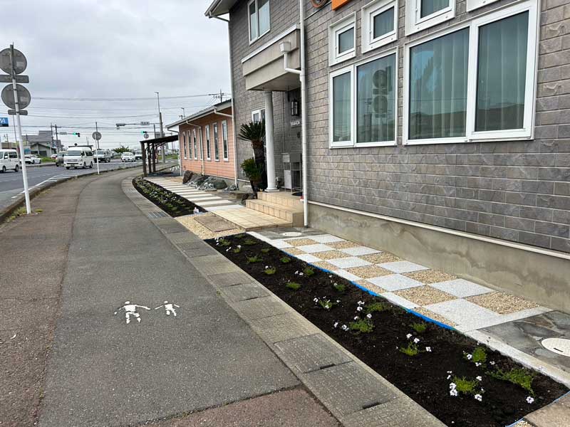 施工後の外構:御影石と真砂土舗装で整えられた石張りのアプローチと、白花のタピアンを用いた植栽が調和する玄関まわり(群馬県玉村町・ダンススクール)